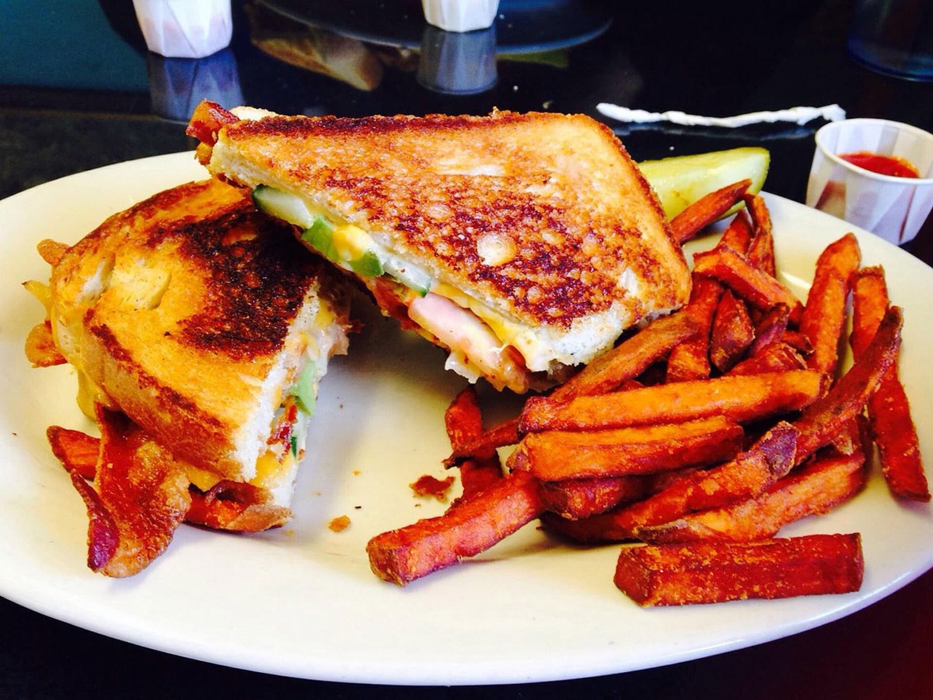 A grilled sandwich with bacon, cheese, and vegetables served with sweet potato fries and a pickle on a white plate.