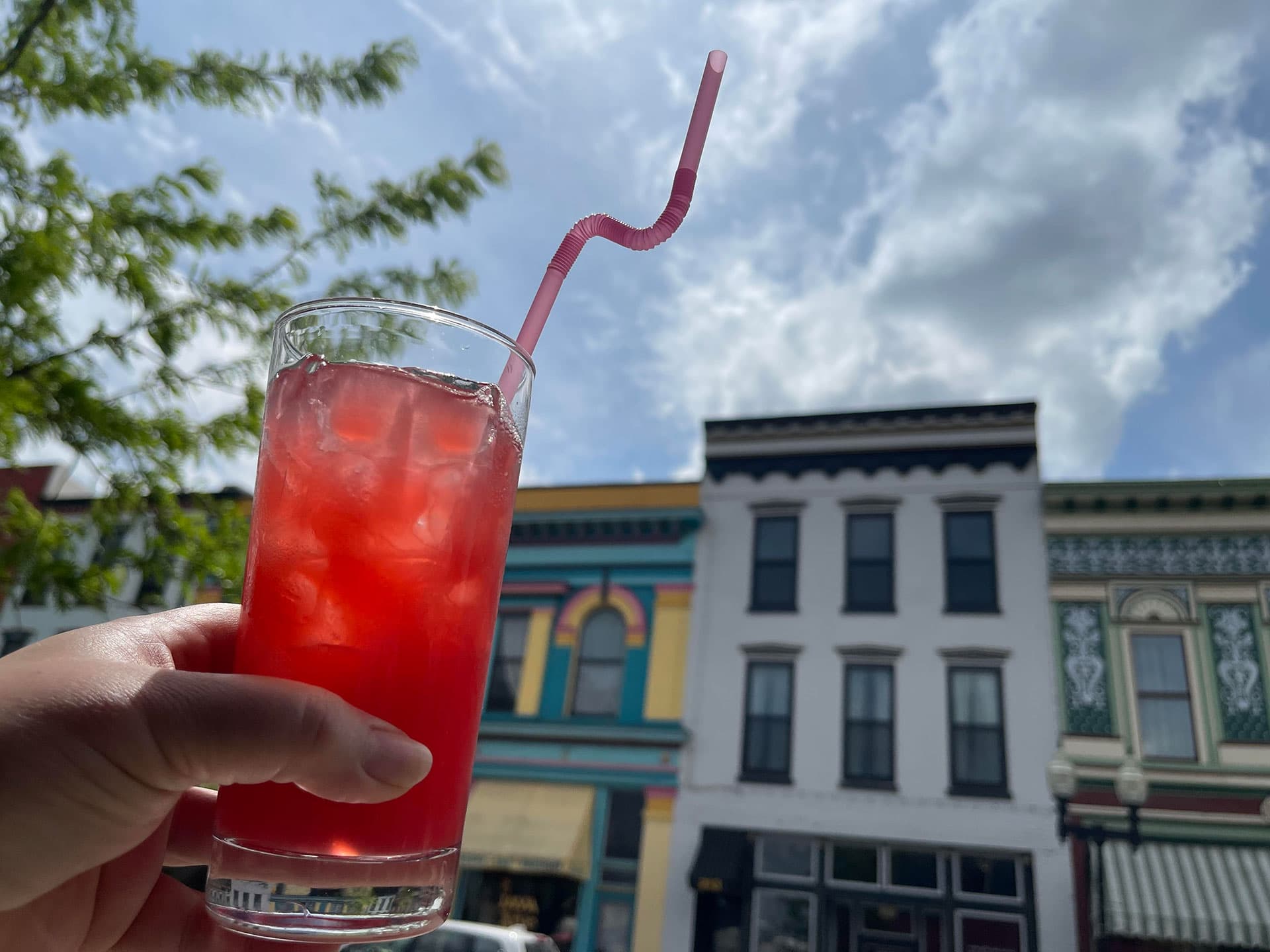 A hand holds a glass of brightly colored beverage with ice against a backdrop of colorful buildings and a blue sky.