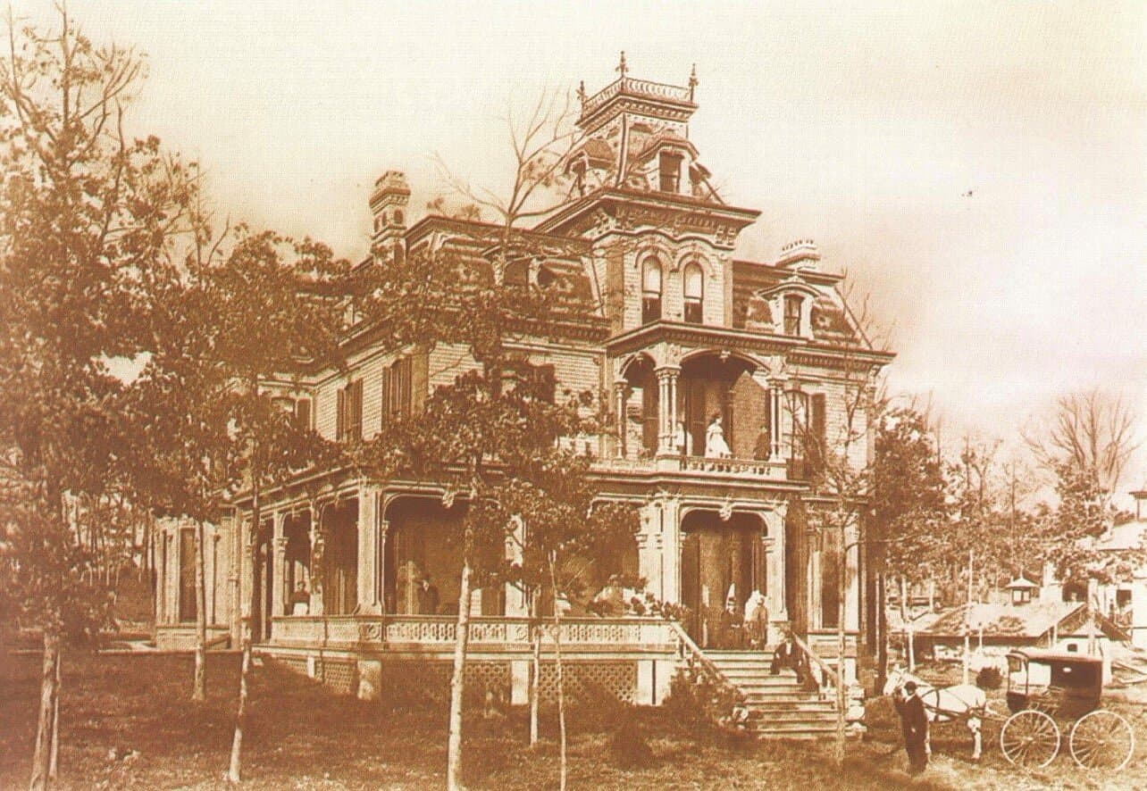 A historical sepia-toned photograph of a large Victorian mansion surrounded by trees, featuring several people on the porch and grounds.