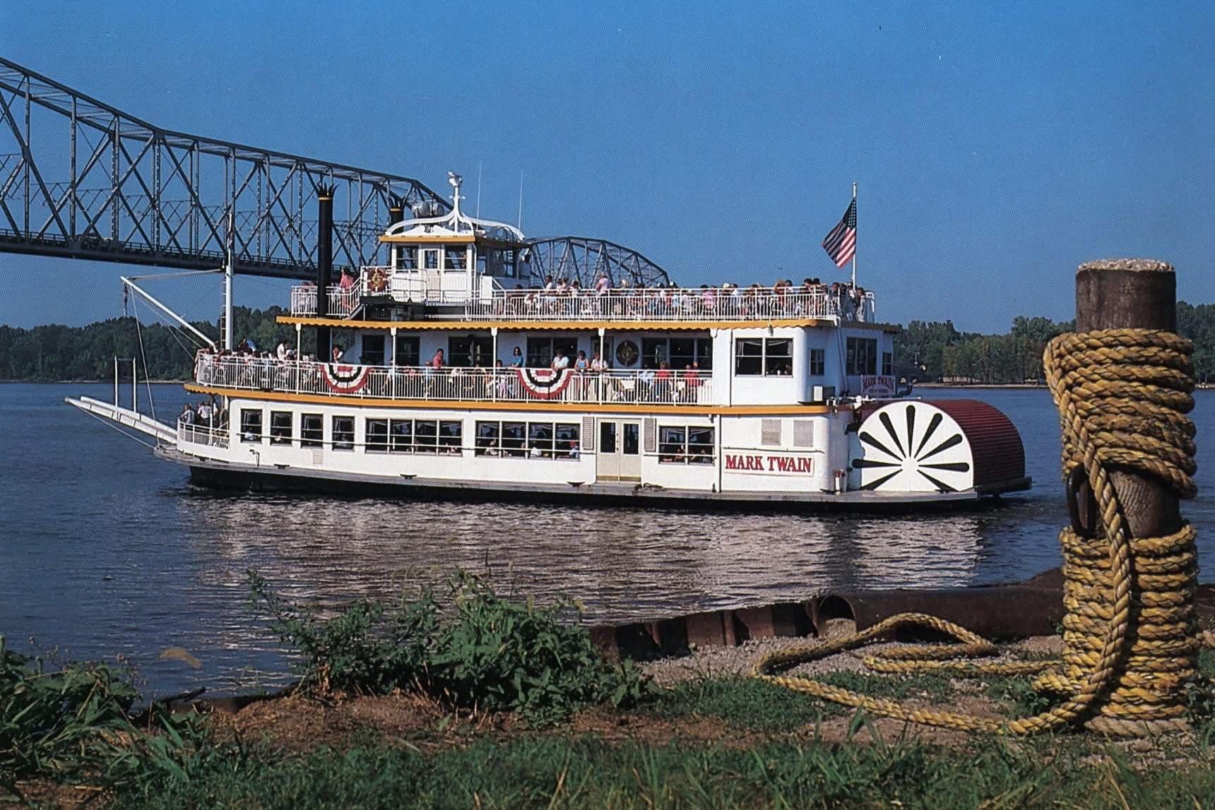 A riverboat named "Mark Twain" sails near a bridge on a clear day.