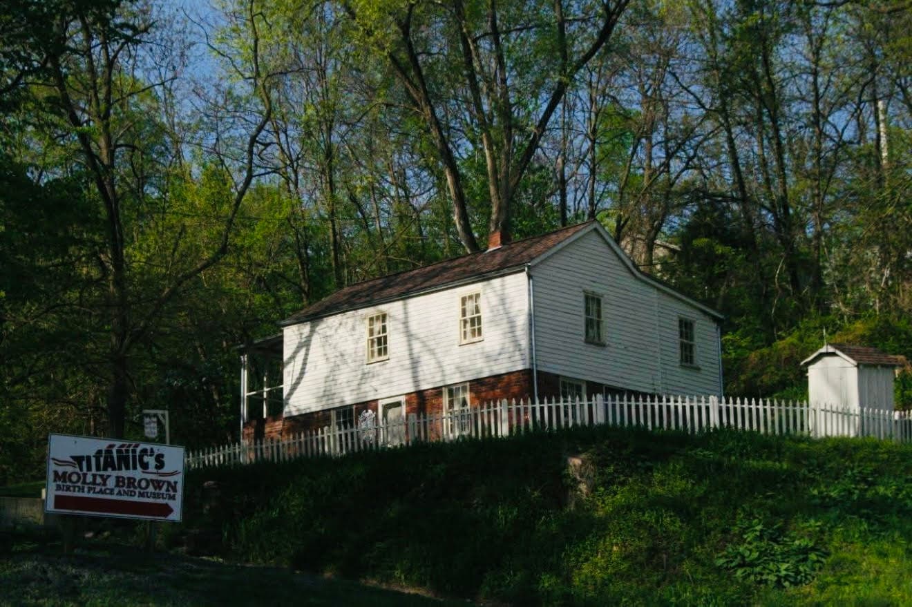 A historic white two-story house with a brick base and a white picket fence, surrounded by trees.