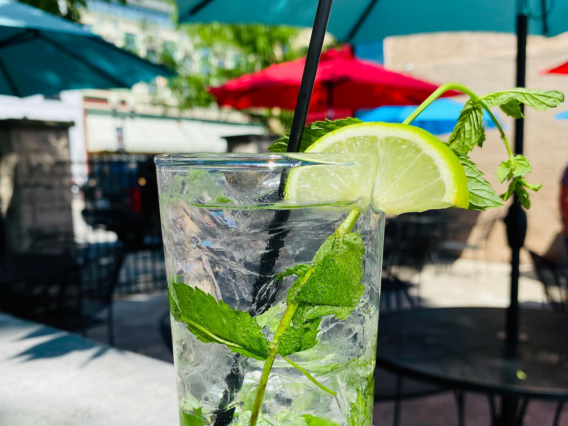 A refreshing mojito with mint and a lime slice in a clear glass, set against a background of colorful patio umbrellas.
