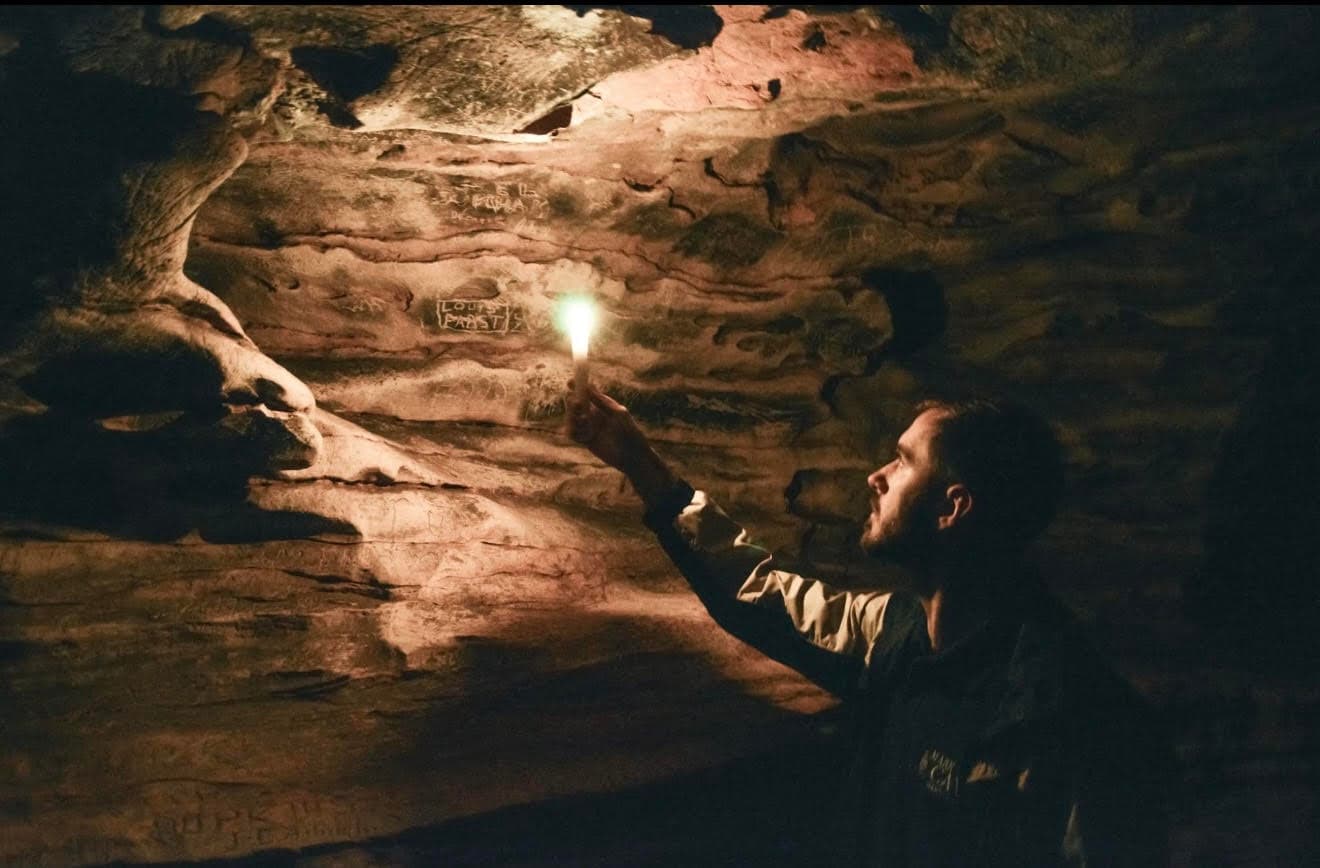 A person explores a dimly lit cave, illuminating ancient carvings with a flashlight.