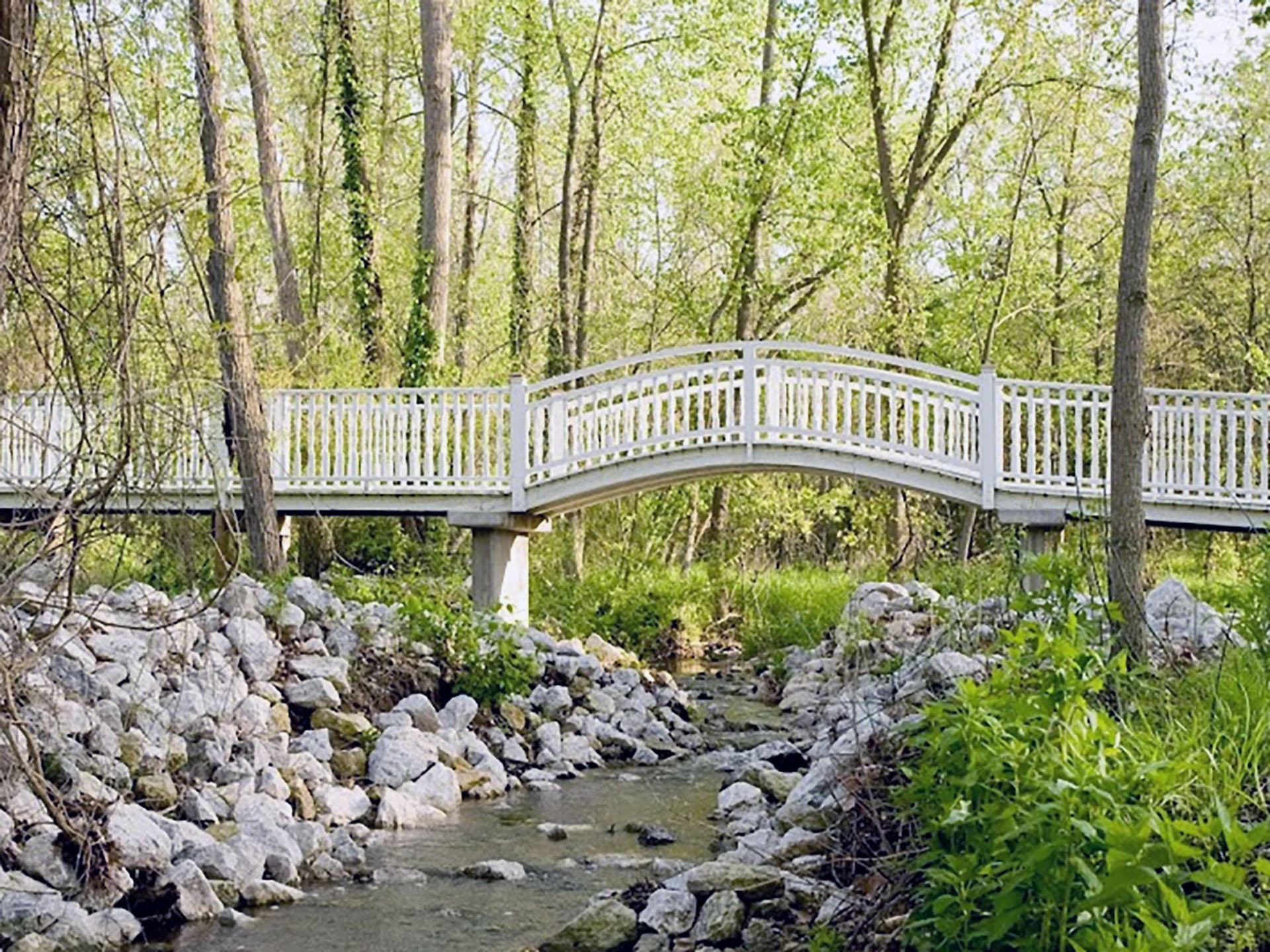 A white wooden bridge arches over a stream surrounded by green trees and rocks.