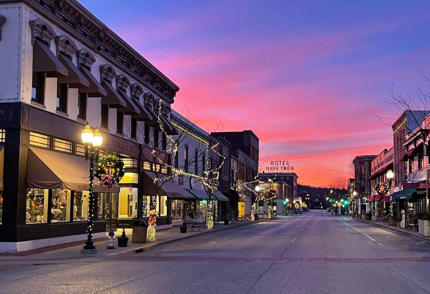 A picturesque street at dusk, adorned with holiday lights and a vibrant sunset backdrop.